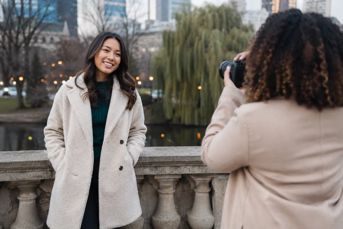 A woman in a light seasonal coat poses by a pond while a photographer captures a fresh profile portrait.